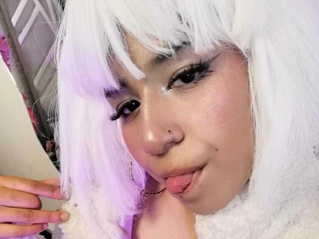 DulceSweett hat weißes Haar, einen Nasenring und steckt ihre Zunge heraus, wobei ihre Augen stark geschminkt sind. Sie trägt eine flauschige weiße Halskrause. Ihre Hand ist in der Nähe ihres Gesichts..