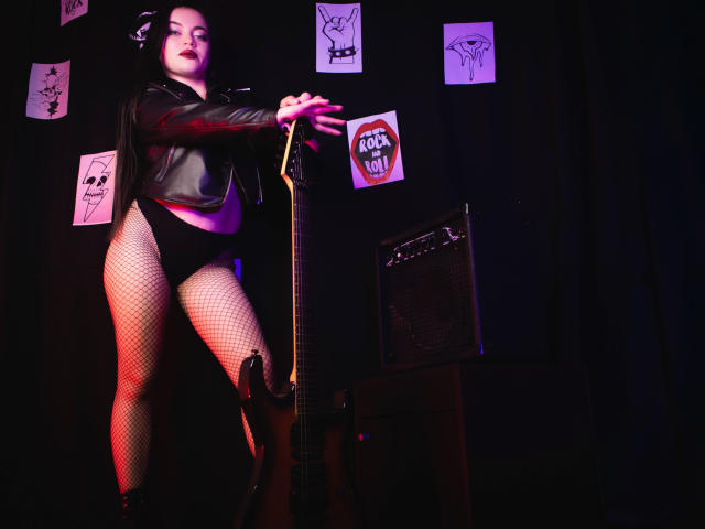 ZoeyStone with pale skin, dark hair, and red lipstick, wearing a black leather jacket, black panties, and fishnet stockings, holding a guitar, standing in front of an amp.