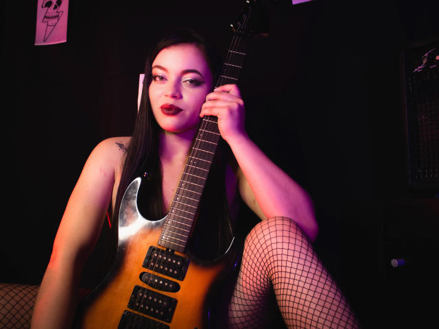 ZoeyStone with long black hair, red lipstick, and fishnet stockings, holding a sunburst electric guitar, sitting with legs apart, showing a tattoo on her right arm.