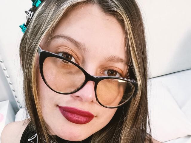 KoraSweet with glasses, red lipstick, and blonde highlights, smiling softly at the camera. Her black-framed glasses are prominent, and she's wearing a black top.