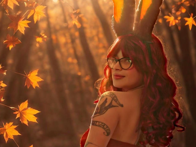 KoraSweet with red curly hair, glasses, bunny ears, and a tattoo on her arm, looking back with a sexy smile, surrounded by orange autumn leaves.