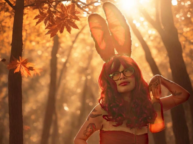KoraSweet with red curly hair, glasses, and bunny ears, holding an orange bottle, showing tattoos, sunbeams, and autumn leaves, standing in a forest.
