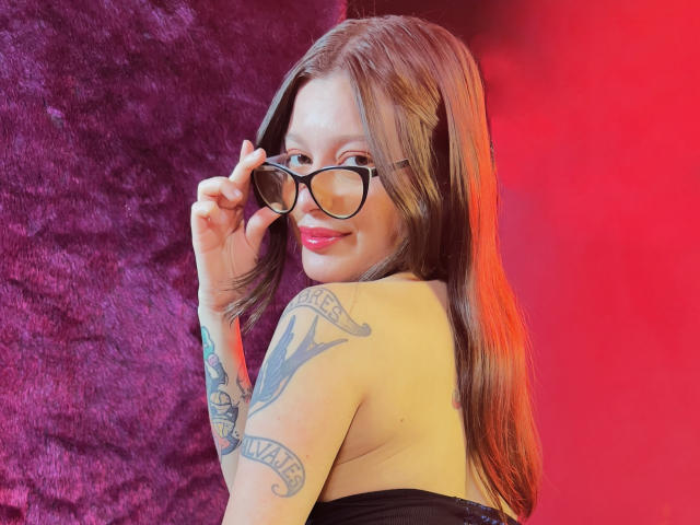 KoraSweet with long brown hair, black glasses, and a smile, showing her tattooed arm, wearing a strapless black top, looking back over her shoulder. Red and purple background.