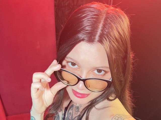 KoraSweet with long brown hair, holding glasses, smiling, showing tattoos on her neck and chest, red lipstick, and a slight nose piercing, in a red-lit photo.