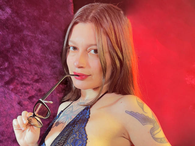 KoraSweet with long brown hair, holding glasses in her mouth, blue lace bra, tattoo on right shoulder, red and purple background, slight smile, fair skin, pink lips.
