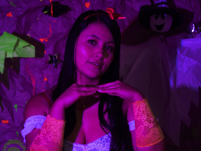 EmilySavage with dark hair, wearing a white lace top, orange lace cuffs, and a red headband with devil horns, smiling with hands near her chin, under purple lighting.