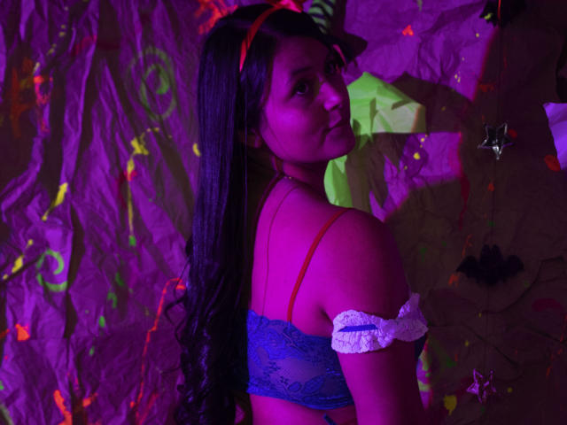 EmilySavage with long black hair, wearing a blue lace bra, red headband, and white off-shoulder ruffle, looking over her shoulder, purple and pink neon lighting.