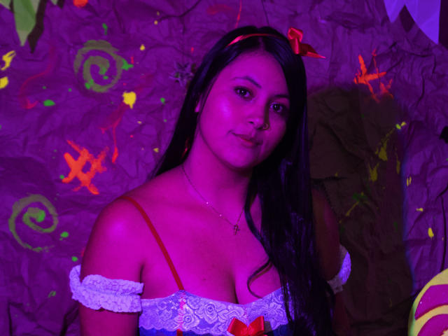 EmilySavage with purple glow, dark hair, red headband, white off-shoulder top showing her tits, necklace, subtle smile, and brown eyes, standing in colorful, neon-lit background.