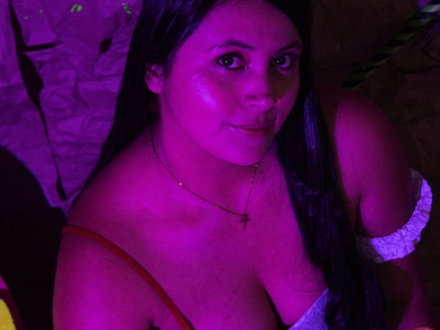 EmilySavage with long black hair, pink-lit skin, wearing a strapless top, showing her large tits, and a cross necklace, smiling slightly at the camera.