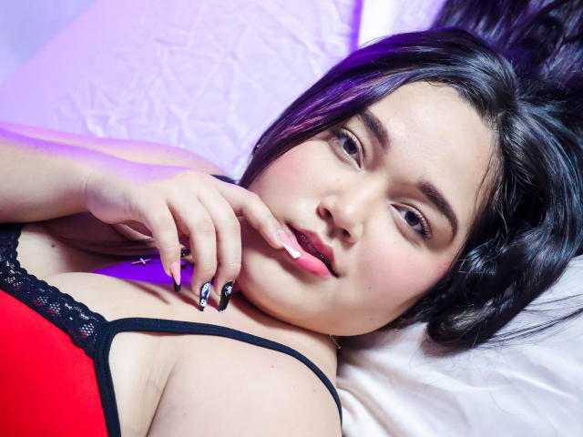 EmilyBrice with black hair, lying on a bed, wearing a red and black lace bra, looking at the camera, finger near her pink, slightly parted lips, with purple lighting.