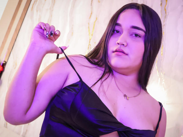 EmilyBrice with long brown hair, purple light, wearing a black satin dress, pulling a strap, showing her chest, black nails, gold necklace, looking at the camera.