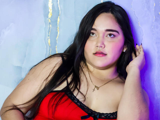 EmilyBrice with long black hair, red and black lace dress, looking to the side, hand in hair, pale skin, pink and blue light on face, necklace with cross.