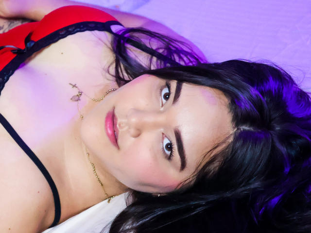 EmilyBrice with black hair, red bra, black lace trim, and gold necklace, lying down, looking at camera with purple lighting, pink lips, and subtle makeup, showing her tits.