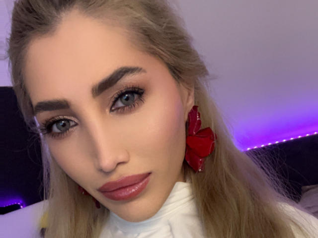 AngelsBack has blue eyes, blonde hair, red bow earrings, and glossy pink lips. Her white turtleneck contrasts with her fair skin and thick, well-groomed eyebrows.