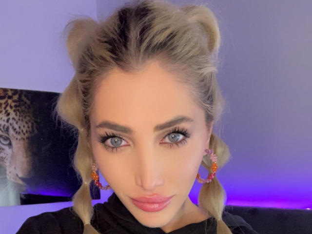 AngelsBack has blonde pigtails, blue eyes, and pink lipstick. She wears colorful earrings and a black shirt, with purple lighting in the background. Her tits are not visible.