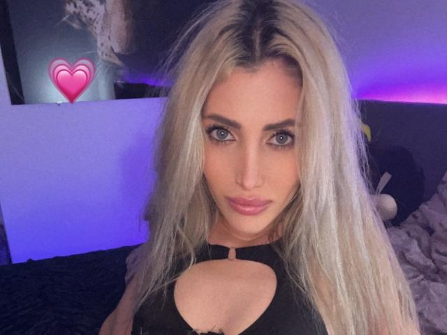 AngelsBack with long blonde hair, blue eyes, and pink lips, wearing a black top with a cutout, showing her tits. A pink heart emoji is on the left.