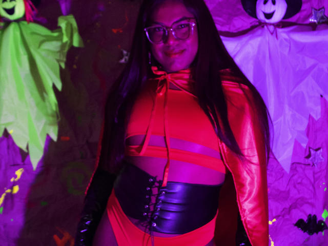 KimmySolange with glasses, long brown hair, red and black latex outfit, corset, and cape, standing, purple lighting, Halloween decorations, green ghost, white ghost in background.