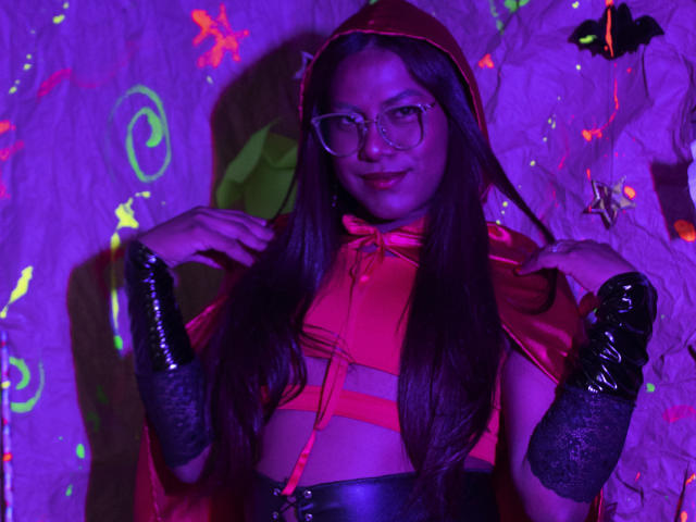 KimmySolange with long brown hair, red hood, glasses, red top, black gloves, and black leather pants, holding her hood up, purple and green neon lights in the background.