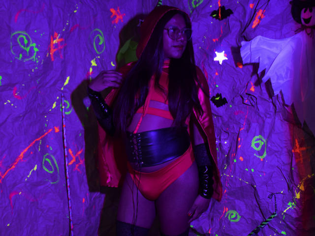KimmySolange in a red hooded costume with glasses, black leather belt, and red underwear, standing with a visible ass, long brown hair, and black arm sleeves, under purple lights.
