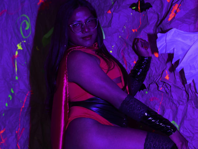 KimmySolange with long dark hair, glasses, red and black latex outfit, black lace gloves, and red cape, lying down, purple and neon lights, sexy, confident, dark skin.