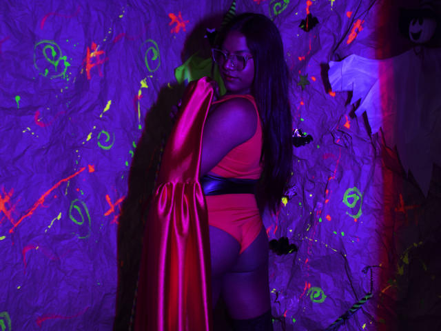 KimmySolange with dark skin, long black hair, glasses, red one-piece, black belt, and pink satin robe, showing her ass, in purple and green neon-lit room.