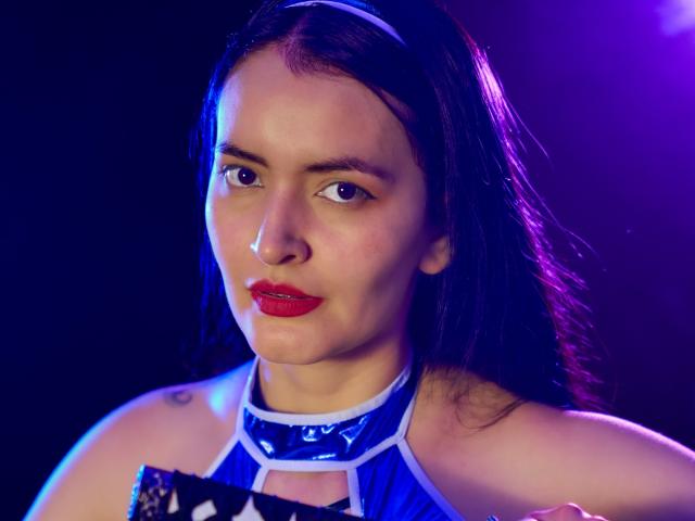 AriaBonnet with red lipstick, blue glossy top, and white headband, showing a small tattoo on her right shoulder, under purple and blue lighting.