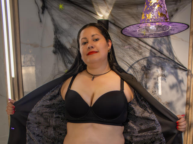 MarianaSmitt se tient devant un miroir, exhibant son soutien-gorge noir et écartant sa veste, dévoilant ses seins et une doublure en maille noire, avec un chapeau violet sur la tête..