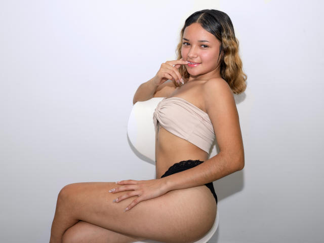 VioletaWilliams with light brown skin, small tits, and black panties, sits on a white chair, smiling, biting her finger, wearing a beige wrap, blonde ombre hair.