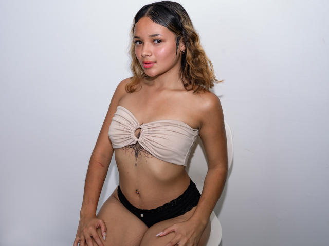 Violeta Williams, con cabello castaño ondulado, un top marrón sin tirantes, ropa interior de encaje negra, un tatuaje pequeño debajo de los senos, sentada en una silla blanca, con una sonrisa sutil, piel clara y complexión delgada..