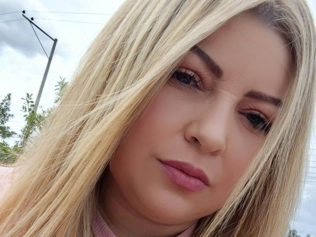 NatalyHami has blonde hair, pink lips, and subtle makeup with brown eyeliner. Her face is slightly down, showing a relaxed, pouty expression. Her nose is centered.