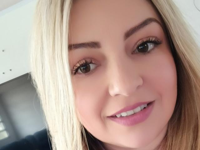 NatalyHami has blonde hair, brown eyes, and is smiling with pink lipstick. Her tits are not visible. She has long eyelashes and a cute face.