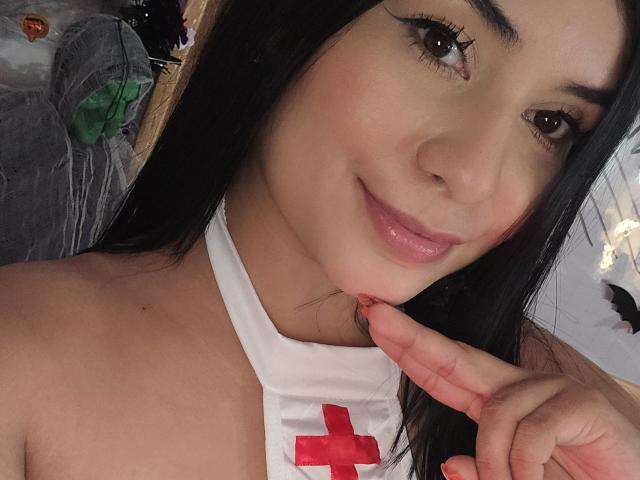 AliceNova with tan skin, black hair, and pink lips, wearing a white nurse costume with a red cross, gently biting her finger, smiling softly, showing her dark eyeliner.