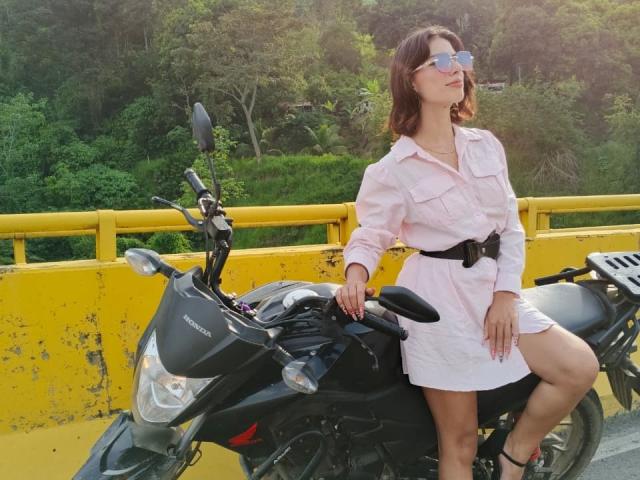 LuisaNinaSky, vêtue d'une robe rose, d'une ceinture noire et portant des lunettes de soleil, assise sur une moto noire, avec une rambarde jaune, des arbres verts et du soleil en arrière-plan. Les seins sont légèrement visibles sous la robe..