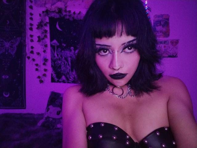 DollyBones with dark makeup, black lipstick, and a black leather strap top with studs, wearing a metal choker, in purple-lit room, showing her tits and a nose ring.