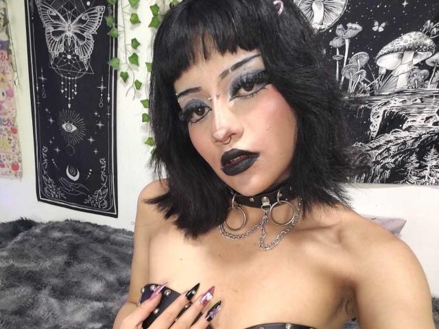 DollyBones with black bob, heavy eyeliner, black lipstick, nose ring, choker with rings, chains, black strapless top, holding boobs, purple nails, looking at camera, dark makeup.