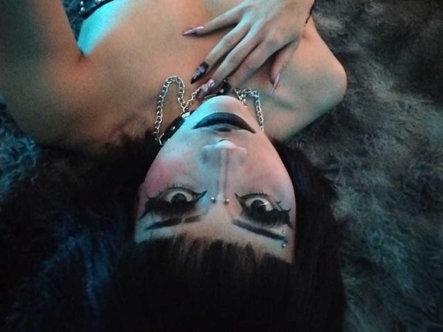 DollyBones with black makeup, chain necklace, and long eyelashes, lying on a furry surface, looking up with a hand on her chest, showing her bare tits.