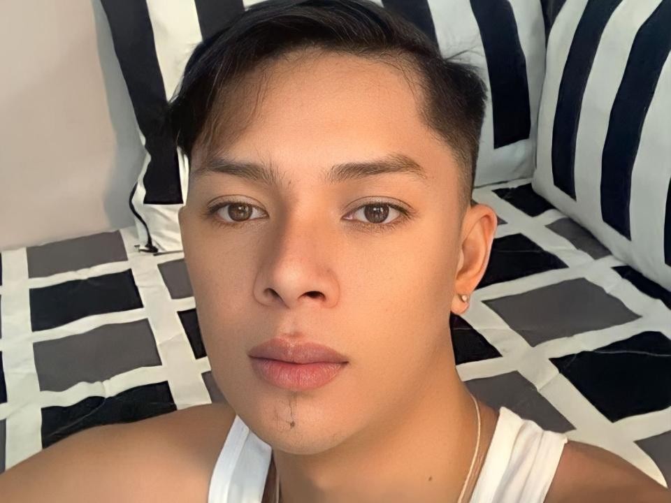 urpinoyasianx con cabello negro corto, ojos marrones, piel clara, una pequeña barba, una camiseta blanca sin mangas, perforaciones en las orejas y una expresión neutra, sobre una cama con un patrón en blanco y negro..