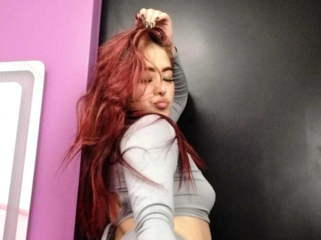 SamaraCuttie stands against a black wall, her long, wavy red hair cascading over her right shoulder. She wears a light gray, long-sleeve crop top, exposing her midriff. Her right hand is raised, fingers tangled in her hair. She has full lips, slightly puckered, and dark, defined eyebrows. Her left arm is slightly bent, adding a casual, confident posture. The wall on the left is purple, with a white-framed mirror partially visible. The composition emphasizes her hair and upper body.