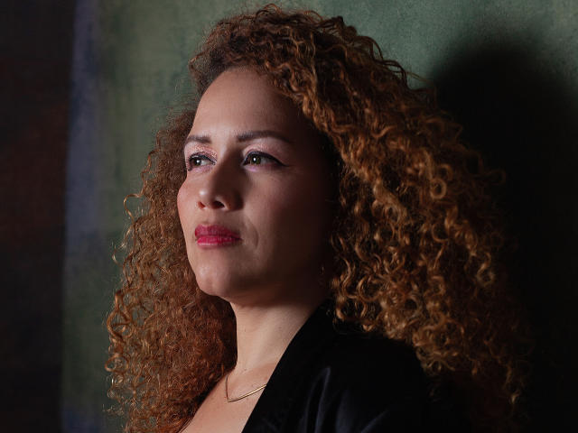 Close-up of RisitosDeoro with curly brown hair, light brown skin, and pink lipstick. She's facing slightly to the right, looking upwards with a subtle smile. She's wearing a black shirt and a thin gold necklace. The lighting is soft, highlighting her curly hair and smooth skin. The background is dark green, creating a contrast with her hair and shirt. RisitosDeoro's makeup includes winged eyeliner, adding a touch of elegance. The overall composition is centered and focused on her upper body and face.