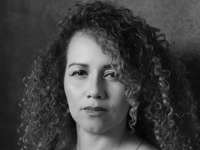 Black and white portrait of RisitosDeoro with curly, voluminous hair. RisitosDeoro's face is centered, showing a neutral expression with slightly parted lips. Thick, dark eyebrows and long eyelashes are visible. Light and shadow play across her face, highlighting her smooth skin and subtle makeup. RisitosDeoro's right eye is partially closed, adding a sense of depth. The texture of her curly hair contrasts with the smoothness of her skin. Simple, dark background.