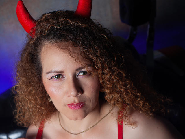 Close-up of RisitosDeoro with curly brown hair wearing red devil horns. RisitosDeoro has light brown skin, green eyes, and pink lipstick. They are wearing a red strap top and a thin gold necklace. The face has a slightly pouty expression. The background is dark with blue light on the right. The image has a realistic, slightly textured appearance. RisitosDeoro's curly hair is thick and voluminous, with the horns adding a playful, demonic element. The light highlights their facial features and hair texture.
