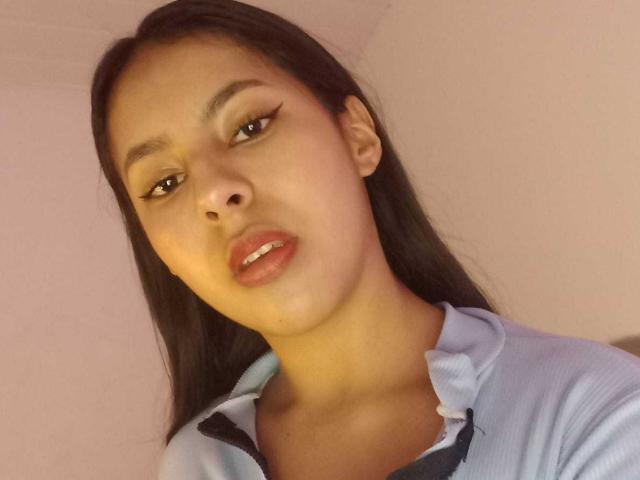 Close-up of SoffiRousee, a young woman with long black hair, light brown skin, and subtle makeup. She has full lips with a slight part and visible teeth. She wears a light blue shirt with a black collar. Her eyebrows are dark and slightly arched. The lighting is soft, highlighting her smooth skin. The composition focuses on her upper body and face. Her expression is neutral, slightly looking to the side. The image has a casual, slightly angled perspective.