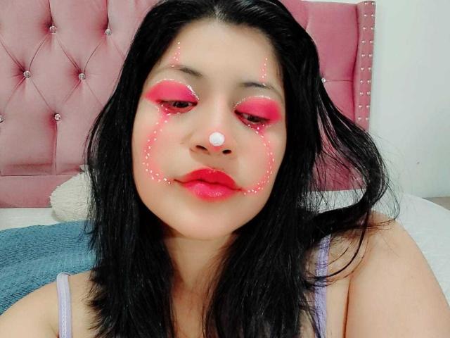 LeylaMiller has pink lipstick, black hair, and pink face makeup with white nose dot, wearing a purple top, sitting against a pink tufted headboard. Her ass is not visible.
