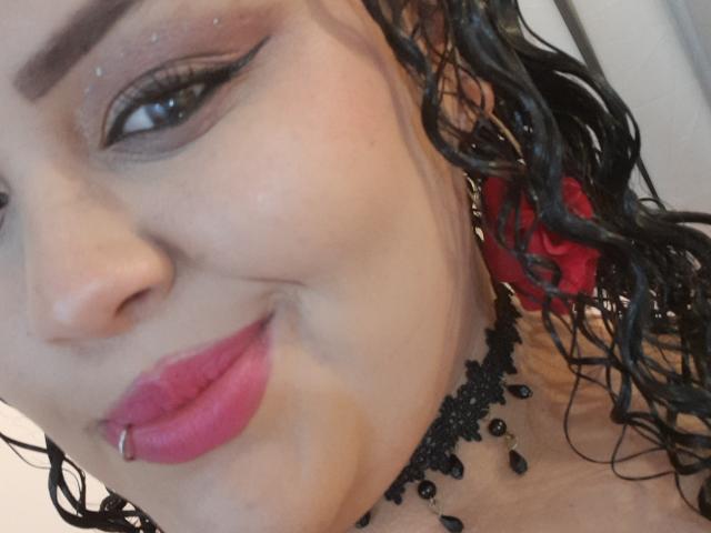 MelannieCarter's face with curly black hair, pink lipstick, black eyeliner, bead necklace, and nose ring, smiling slightly. Her skin is light, and she has a pink lip piercing.