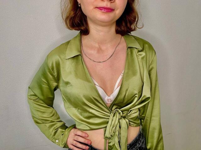 Steisii has pale skin, red hair, and pink lipstick. She wears a green satin shirt tied at the front, exposing her small tits in a white bra.
