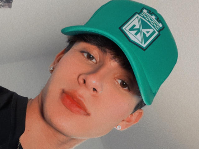 Angelitocute has a green cap with a white logo, wearing earrings, and a black shirt, with a slightly tilted head and a pouty face. Brown eyes, medium skin tone.