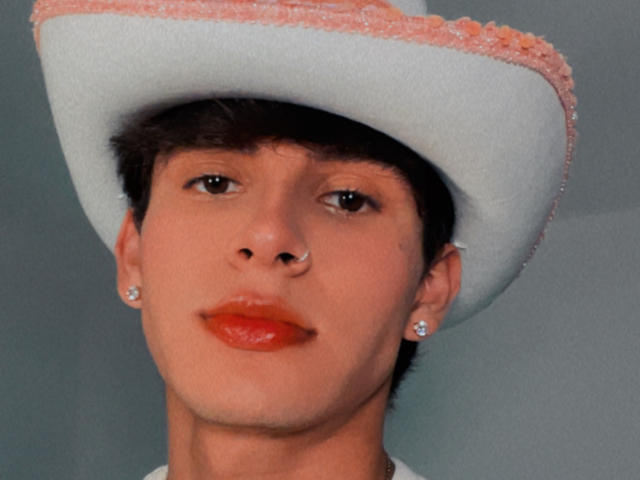 Angelitocute has a white cowboy hat with pink trim, short brown hair, nose ring, red lipstick, and diamond earrings, looking straight at the camera.