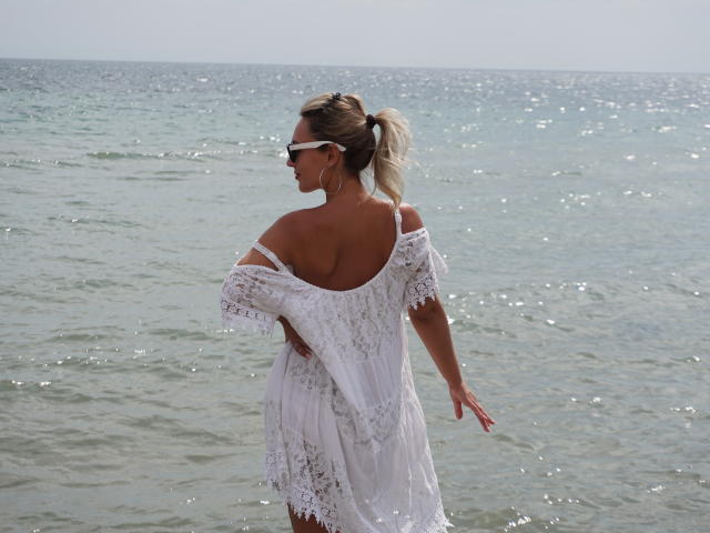 MillaXQueen with white lace dress, off-shoulder, showing her ass, standing in water, blonde ponytail, sunglasses, looking back, clear sky, ocean background, sunlight, subtle waves.
