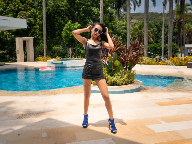 EvelinLuna, vêtue d'une robe noire moulante et de talons bleus, se tient debout, les mains dans les cheveux, montrant ses fesses et ses petites seins, près d'une piscine, avec des lunettes de soleil, en plein jour..