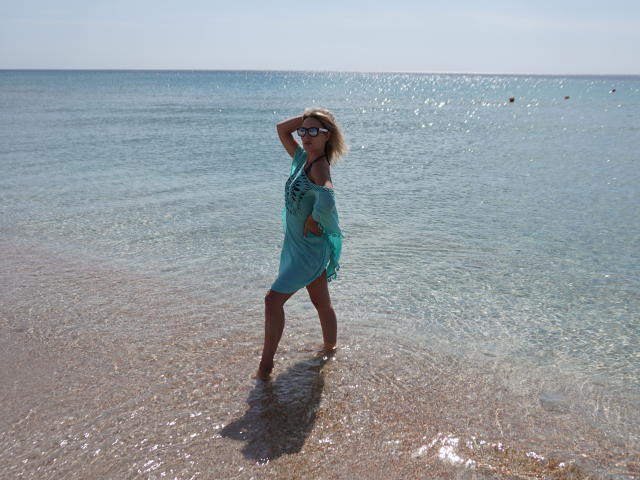 MillaXQueen in a teal dress standing in shallow water, hands in her blonde hair, wearing sunglasses, with her ass and small tits slightly visible, under a clear blue sky.